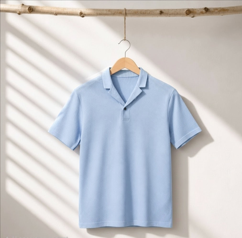 A light blue short-sleeve polo shirt hanging on a wooden hanger from a rustic branch, set against a warm off-white wall with soft diagonal shadows.