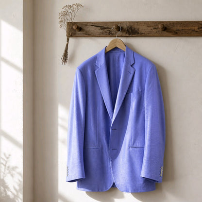 A light blue textured blazer jacket hanging on a wooden hanger from a rustic wooden wall rack, with soft sunlight casting shadows on the left against a textured off-white wall.