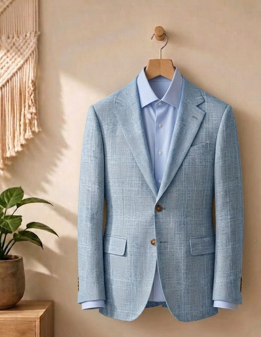 A light blue textured blazer jacket styled over a matching light blue dress shirt, hanging on a wooden hanger against a warm beige wall with a macrame wall hanging and potted plant in the background.