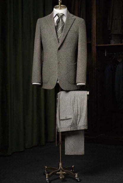 A light brown wool and cashmere single breasted jacket displayed on a mannequin, with pinstripe wool trousers placed below it. The jacket is presented against a green background with a notched lapels design and front pockets.
