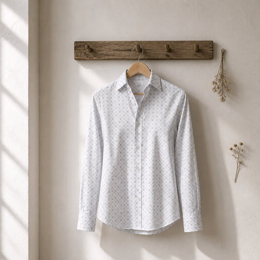 A light gray and white patterned long-sleeve button-up shirt hanging on a wooden hanger from a rustic wooden wall rack, set against a warm beige wall with soft window light shadows.