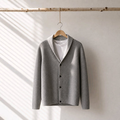A light gray ribbed knit cardigan sweater with a shawl collar, hanging on a wooden hanger from a rustic horizontal branch. The cardigan is layered over a white t-shirt and is photographed against a neutral off-white wall with soft diagonal window shadow patterns.