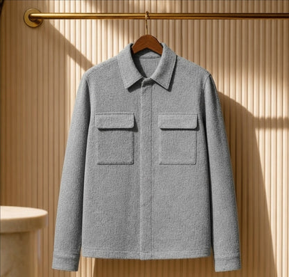 A light gray textured jacket with a pointed collar and two front flap pockets, hanging on a wooden hanger against a beige slatted wall with warm sunlight and shadows.