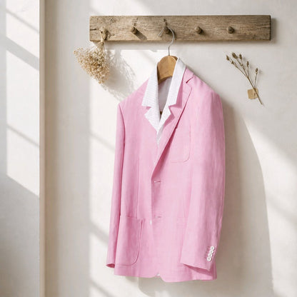 A light pink linen-blend blazer jacket with a white striped inner collar, hanging on a wooden hanger from a rustic wooden peg rack on a sunlit cream wall.