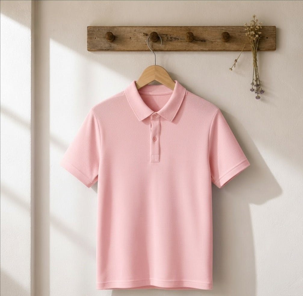 A light pink short-sleeve polo shirt hanging on a wooden hanger from a rustic wooden wall rack with dried flowers, set against a warm off-white wall with soft window light shadows.