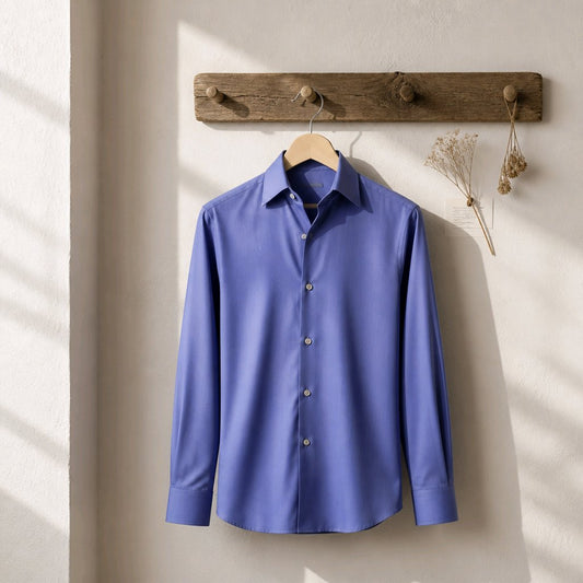 A long-sleeve button-up shirt in a periwinkle blue color hangs on a wooden hanger from a rustic wooden peg rack mounted on a textured off-white wall.