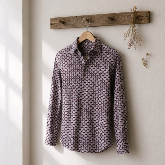 A long-sleeve button-up shirt with a small repeating geometric pattern in dark brown and muted purple, hanging on a wooden hanger from a rustic wooden peg on a textured off-white wall.