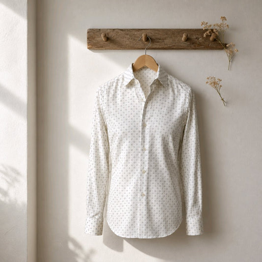 A long-sleeve button-up shirt with a small white geometric pattern on a cream base, hanging on a wooden hanger from a rustic wooden coat rack against a textured off-white wall.