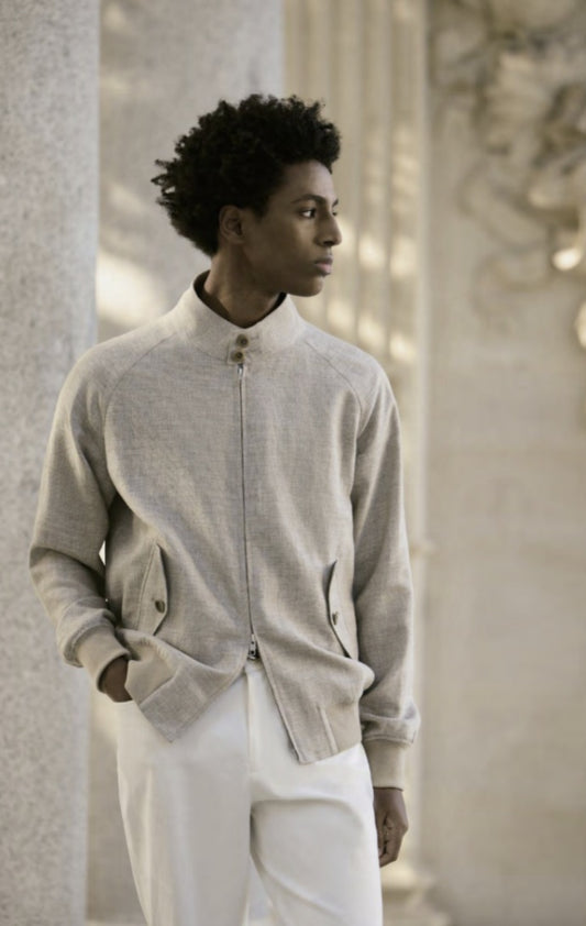 A male model wearing a beige wool & linen-silk & spandex jacket with white trousers. The jacket features a Nehru collar, side flapped button pockets, and an elastic hem.