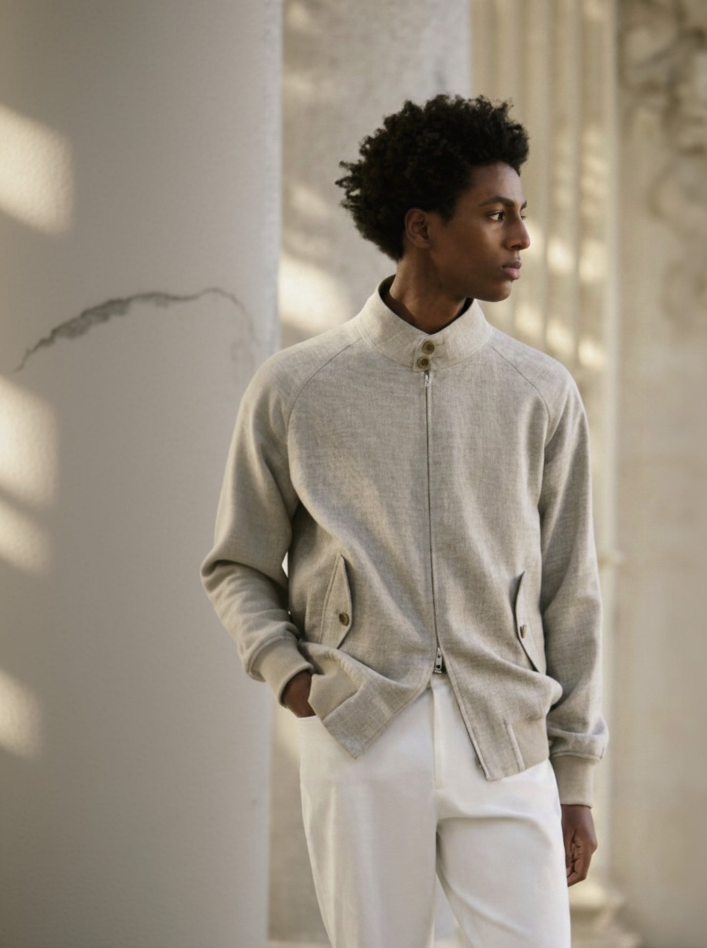 A male model wearing a beige wool & linen-silk & spandex jacket with white trousers. The jacket features a Nehru collar, side flapped button pockets, and an elastic hem.