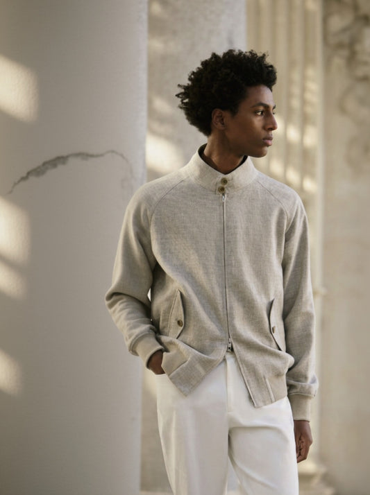 A male model wearing a beige wool & linen-silk & spandex jacket with white trousers. The jacket features a Nehru collar, side flapped button pockets, and an elastic hem.