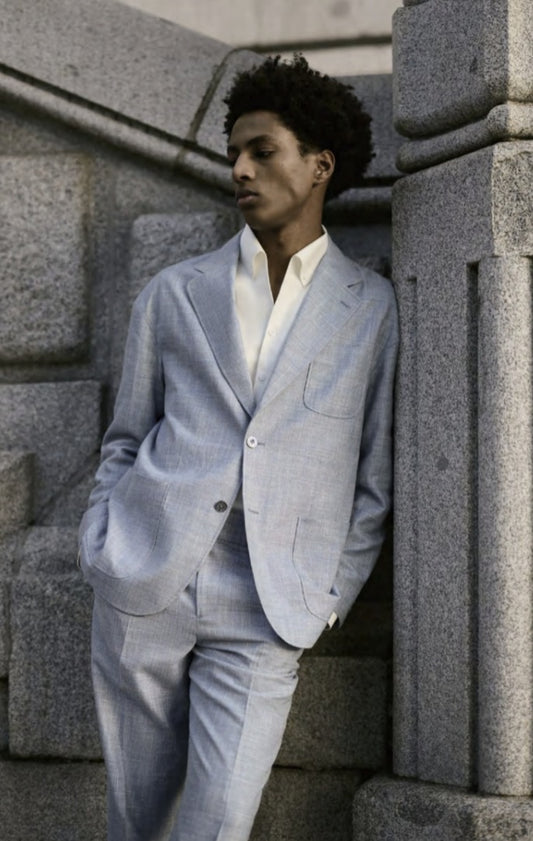 A male model wearing a light blue two-piece suit with a notched lapel, round patch pockets, and a baby blue paisley lining. The suit has a slim fit and the model is standing against a backdrop of stone columns.
