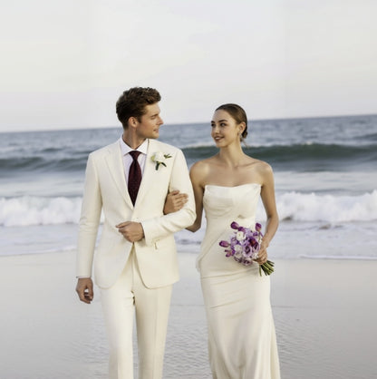 A man and a woman walking on a beach wearing elegant ivory linen suits. The man is in a tuxedo with a notched lapel and the woman is in a dress with a floral design.