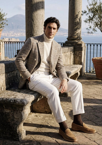A man sits on a stone bench in an outdoor terrace setting with columns and a scenic lake view in the background. He is wearing a brown and beige checkered blazer, a cream turtleneck sweater, white trousers, dark brown socks, and brown suede loafers.