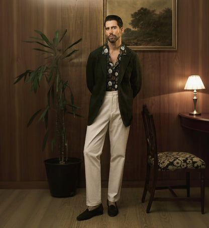A man standing in a room with wooden panel walls, wearing a dark green textured two-piece suit with a notched lapel, patch pockets, and a double vent. The suit has a silver range pattern and appears to be matte finish.