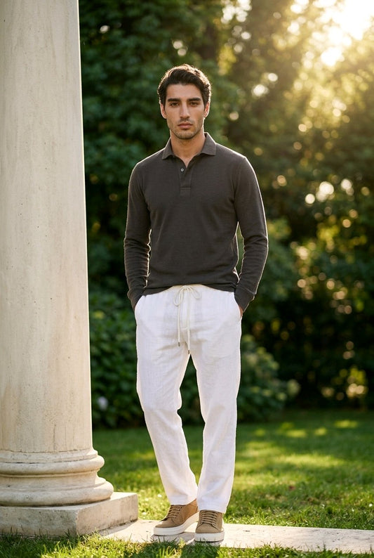 A man standing outdoors next to a large stone column, wearing a dark charcoal long-sleeve polo shirt, white drawstring trousers, and tan sneakers, with a sunlit green garden background.