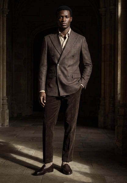 A man stands in a dimly lit stone corridor wearing a tailored double-breasted suit in brown and taupe tones, with a light shirt and dark loafers.