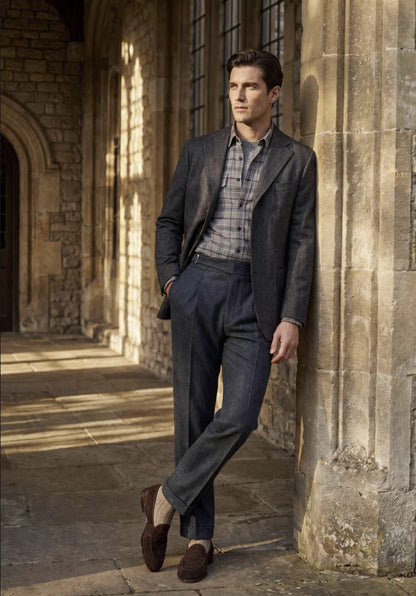 A man stands in a stone corridor wearing a dark charcoal gray suit, a blue and white plaid shirt, brown loafers, and brown socks.