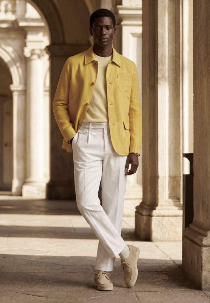 A man stands in a sunlit stone arcade wearing a bright yellow overshirt, cream shirt, white trousers, and beige sneakers.