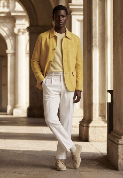 A man stands in a sunlit stone arcade wearing a bright yellow overshirt, cream shirt, white trousers, and beige sneakers.