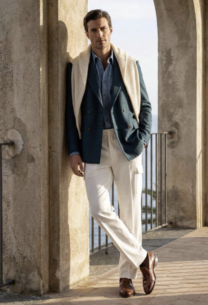 A man stands in an outdoor stone archway wearing a tailored outfit with a navy double-breasted blazer, light blue dress shirt, cream vest, cream trousers, and brown leather shoes.