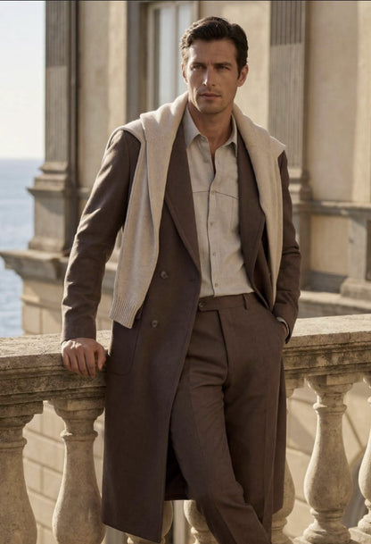 A man stands on a stone balcony wearing a long brown overcoat, matching brown trousers, a light beige shirt, and a cream knit sweater draped over his shoulders, with a coastal building in the background.