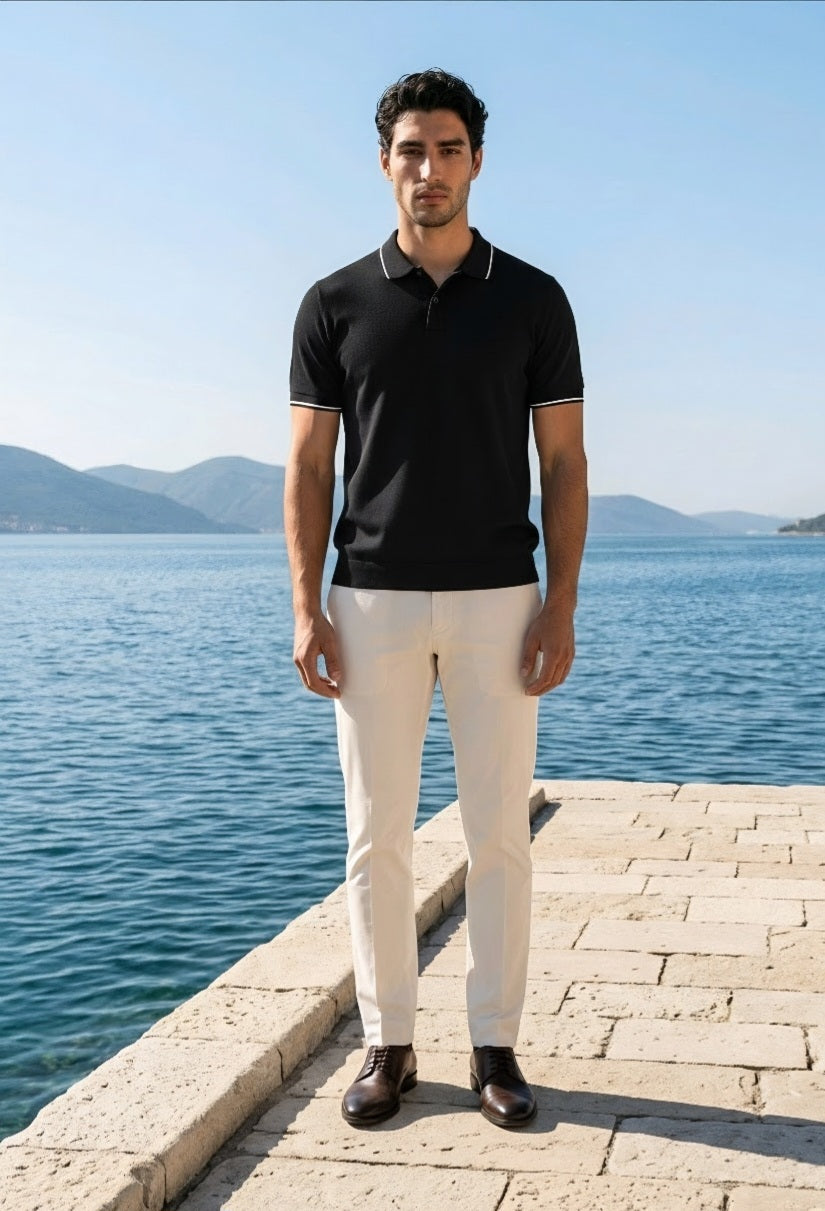 A man stands on a stone pier by the sea wearing a black short-sleeve polo shirt with white piping, off-white trousers, and dark brown shoes.