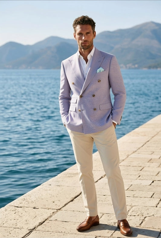 A man stands on a stone pier by the sea wearing a light lavender double-breasted blazer, cream trousers, and brown leather shoes, with mountains and blue water in the background.