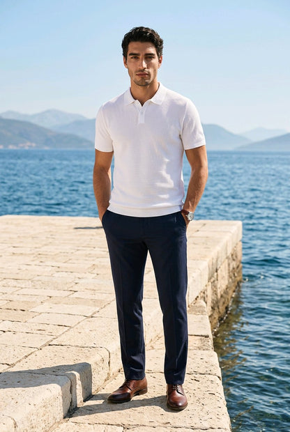 A man stands on a stone pier by the sea wearing a white short-sleeve polo shirt, navy trousers, and brown leather shoes, with blue water and distant mountains in the background.