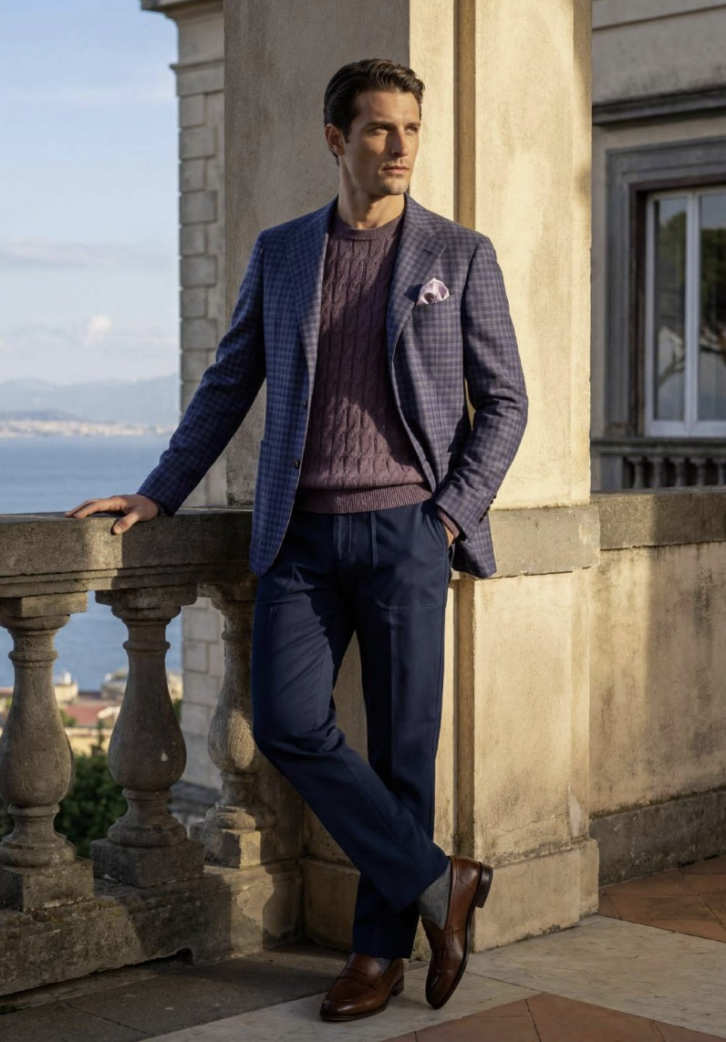 A man stands outdoors against a stone building column, wearing a tailored navy blazer with a subtle windowpane pattern over a mauve cable-knit sweater, navy trousers, and brown dress shoes.