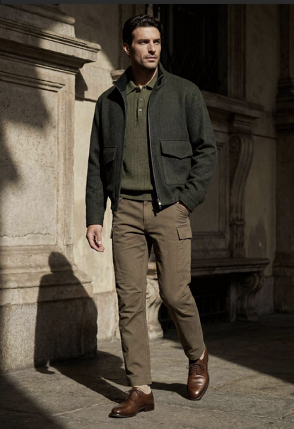 A man stands outdoors against a sunlit stone building, wearing a dark olive green zip-front jacket over a matching green polo sweater, tan cargo pants, and brown leather dress shoes.