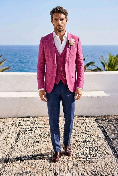 A man stands outdoors by the ocean wearing a bright pink textured blazer and matching vest over a white shirt, with navy trousers and brown shoes. A pink boutonniere is pinned to his jacket, and a white wall and pebble ground are visible behind him.