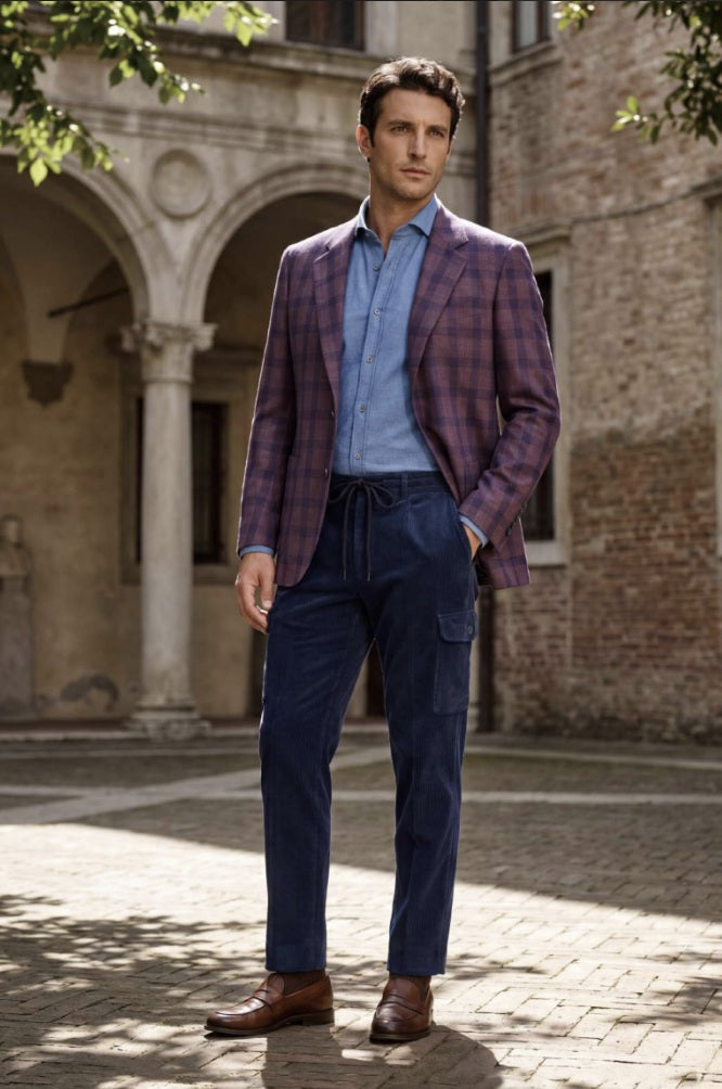 A man stands outdoors in a courtyard wearing a purple and blue plaid blazer, a light blue button-down shirt, dark navy trousers, and brown leather shoes.
