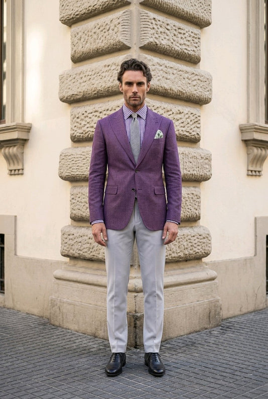 A man stands outdoors in front of a stone building corner wearing a purple blazer, light gray trousers, a light blue patterned shirt, a matching tie, and black dress shoes.