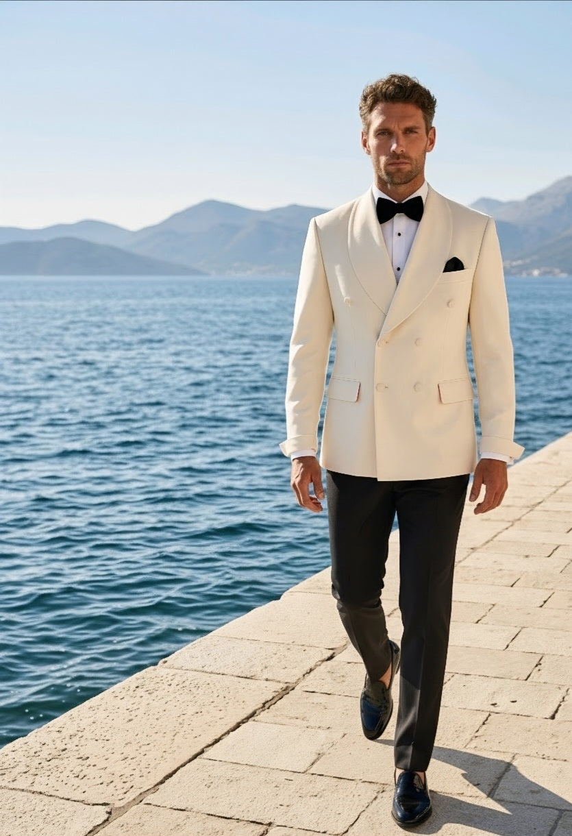 A man walking on a stone pier by the sea wearing a cream double-breasted tuxedo jacket with black trousers, a black bow tie, and a black pocket square, with mountains in the background.