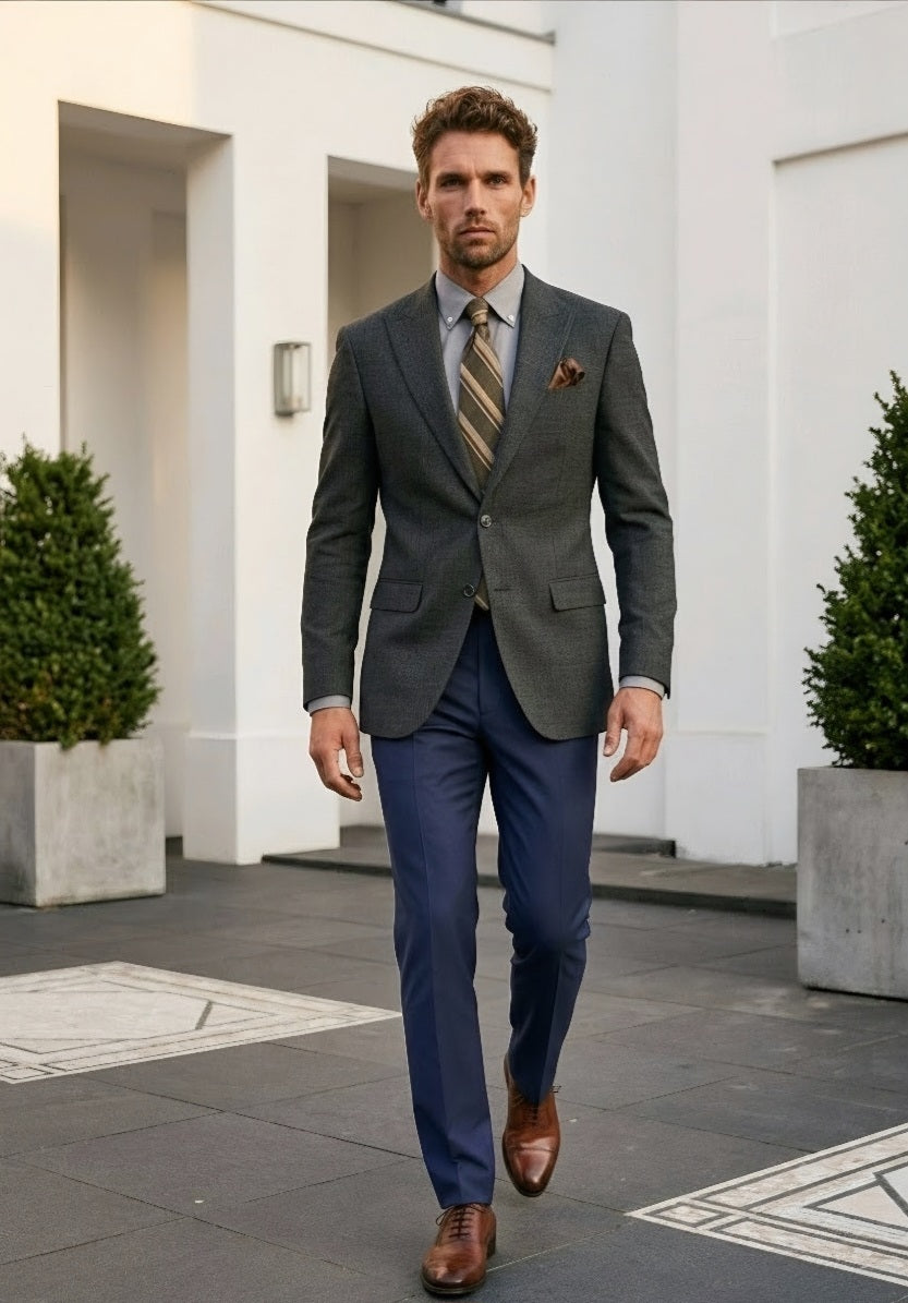 A man walking outdoors in a gray textured blazer, navy blue trousers, a striped tie, and brown leather dress shoes, standing in front of a modern white building.