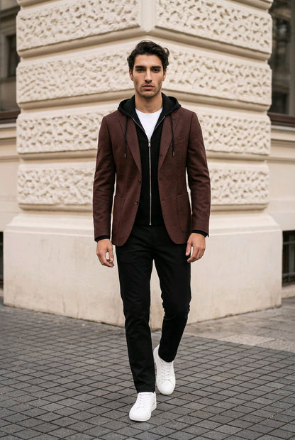 A man walking outdoors in front of a beige stone building, wearing a dark brown blazer over a black hoodie and black pants with white sneakers.