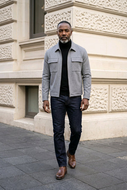 A man walking outdoors in front of a beige stone building, wearing a light gray textured jacket over a black turtleneck, dark blue jeans, and brown leather boots.