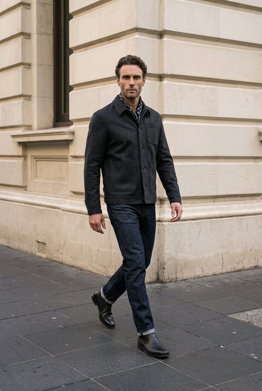 A man walking outdoors on a city sidewalk next to a beige stone building, wearing a dark charcoal jacket, dark blue jeans, and dark boots.