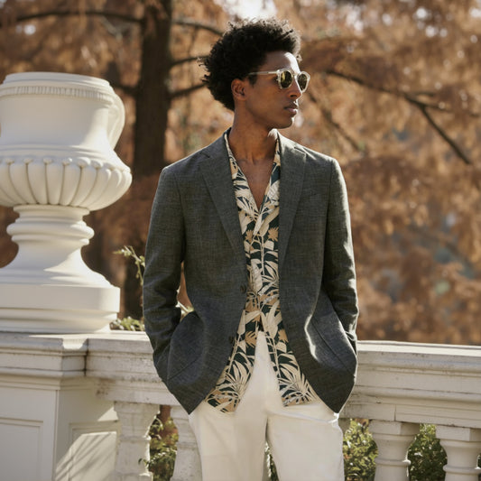 A man wearing a Italian green linen-cotton jacket with a notched lapel and dark brown buttons, paired with white trousers and a white paisley shirt.