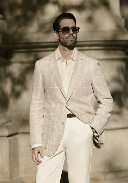 A man wearing a beige and white check linen jacket with white trousers and sunglasses standing in front of a stone wall.