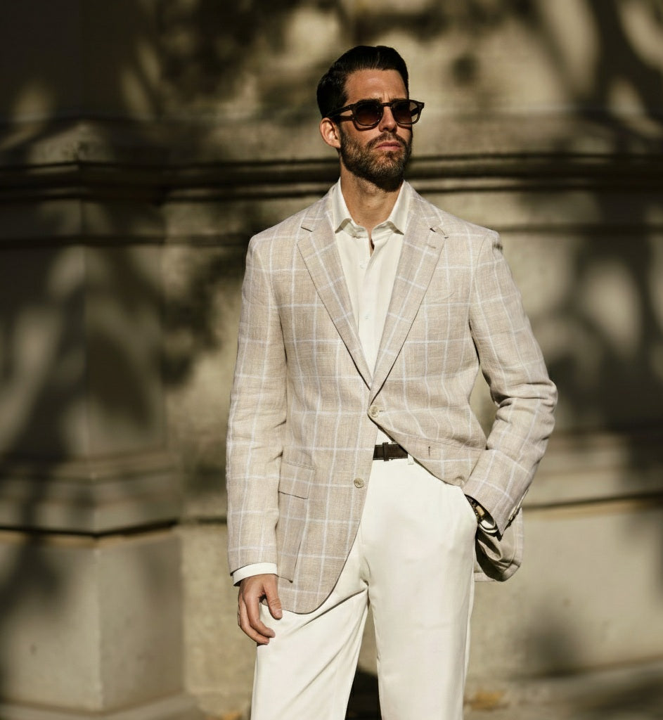A man wearing a beige and white check linen jacket with white trousers and sunglasses standing in front of a stone wall.
