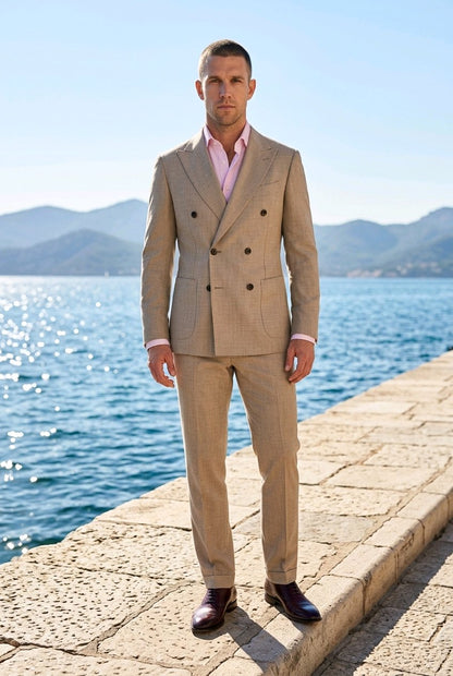 A man wearing a beige double-breasted suit with a pink shirt and dark brown dress shoes, standing on a stone pier with a blue sea and mountains in the background.