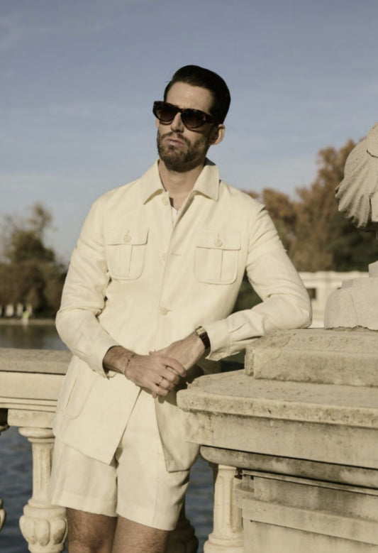 A man wearing a beige two-piece suit with patch pockets, a shirt collar, and sunglasses, standing by a water feature.
