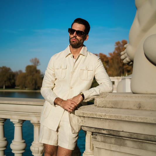 A_man_wearing_a_beige_two-piece_suit_with_patch_pockets_a_shirt_collar_and_sunglasses_standing_by_a_water_feature.