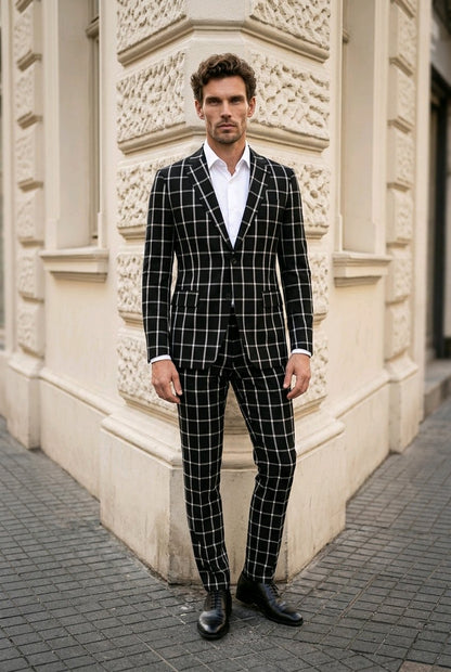 A man wearing a black and white windowpane plaid suit with a white dress shirt and black dress shoes, standing on a paved street in front of a beige stone building.