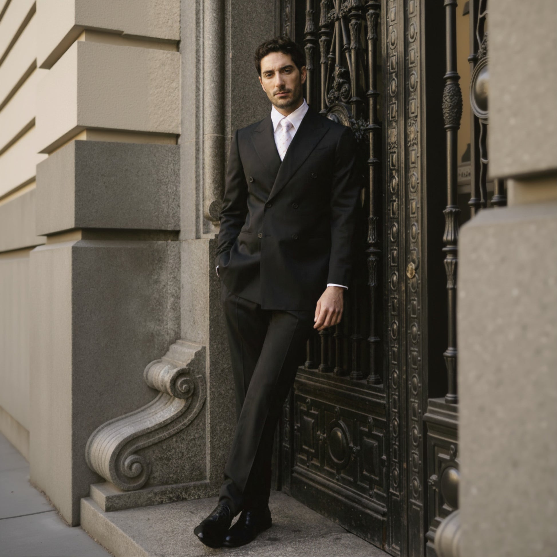 A man wearing a black double-breasted, wool-spandex two-piece suit standing near a decorative wall.