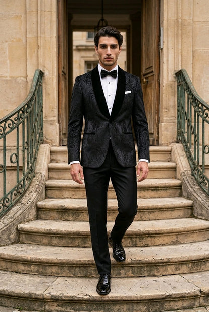 A man wearing a black patterned tuxedo jacket with black velvet shawl lapels, a white shirt, and a black bow tie walks down stone stairs in front of a classical building.