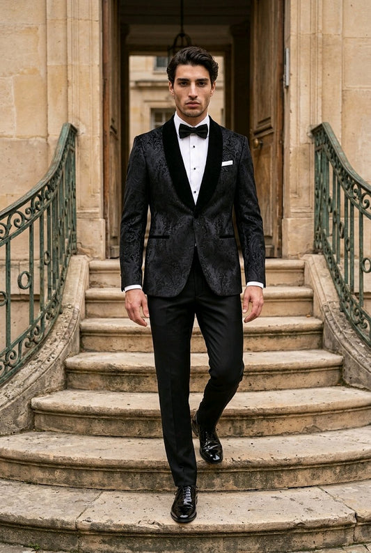 A man wearing a black patterned tuxedo jacket with black velvet shawl lapels, a white shirt, and a black bow tie walks down stone stairs in front of a classical building.