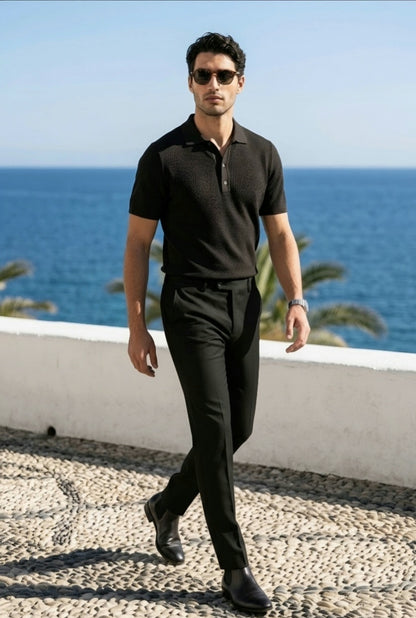 A man wearing a black short-sleeve polo shirt and black trousers walks outdoors by the sea, wearing sunglasses and black dress shoes.